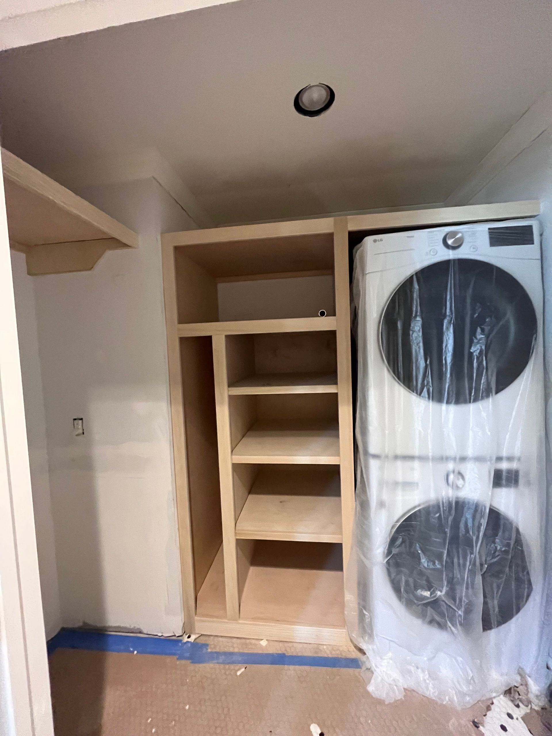 A laundry closet in progress with built-in wooden shelving beside a stacked white washer and dryer unit.