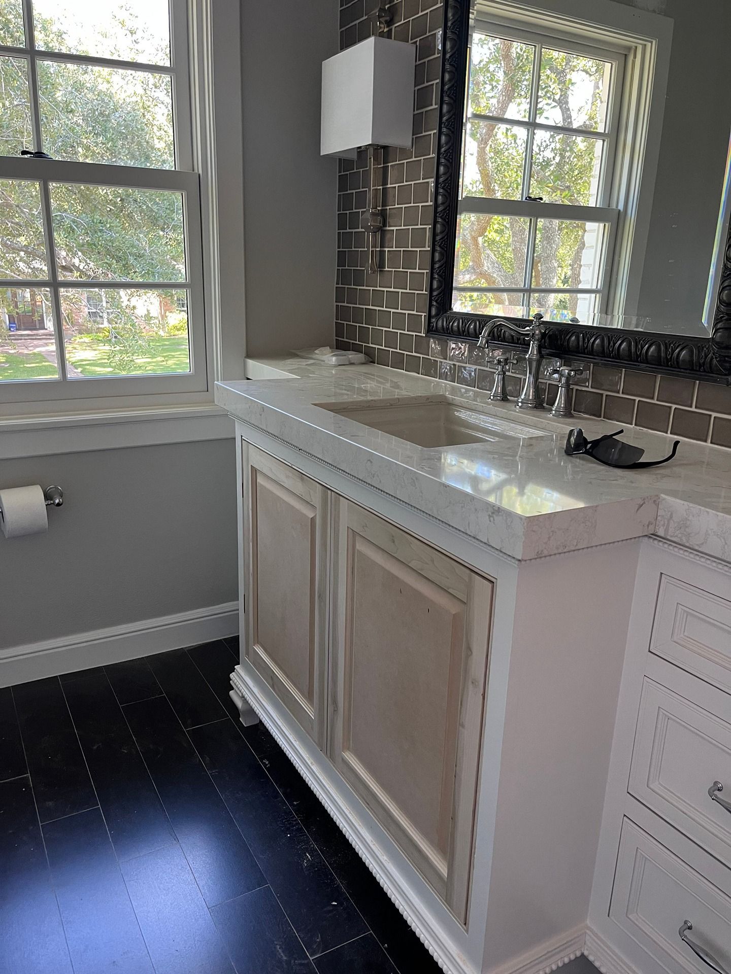 A vanity with a white countertop and natural wood cabinet doors stands in a bathroom with dark floors and brick tiles.