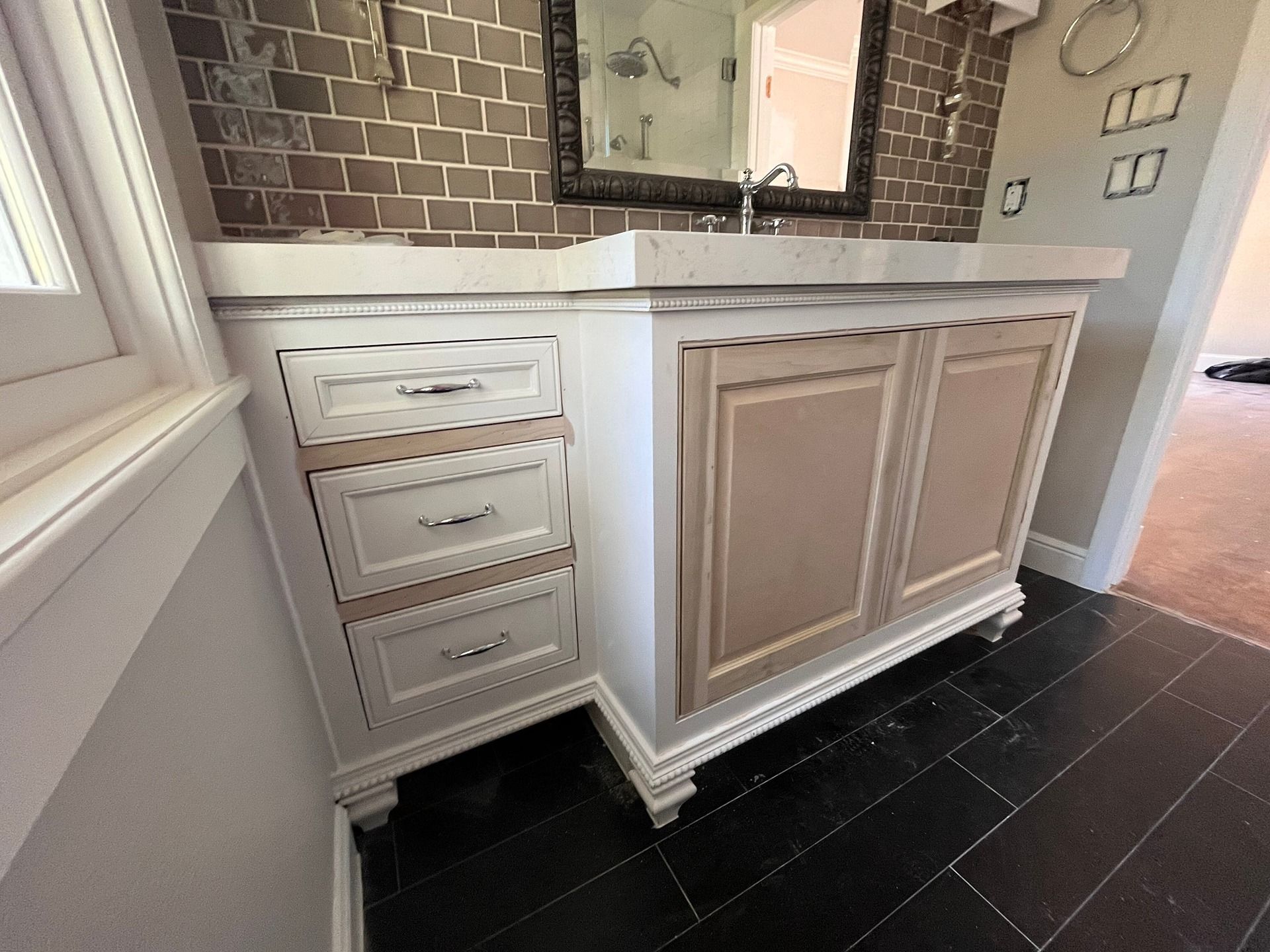 A white bathroom vanity with three drawers and two cabinet doors, set against a brown brick-style wall and black tile.