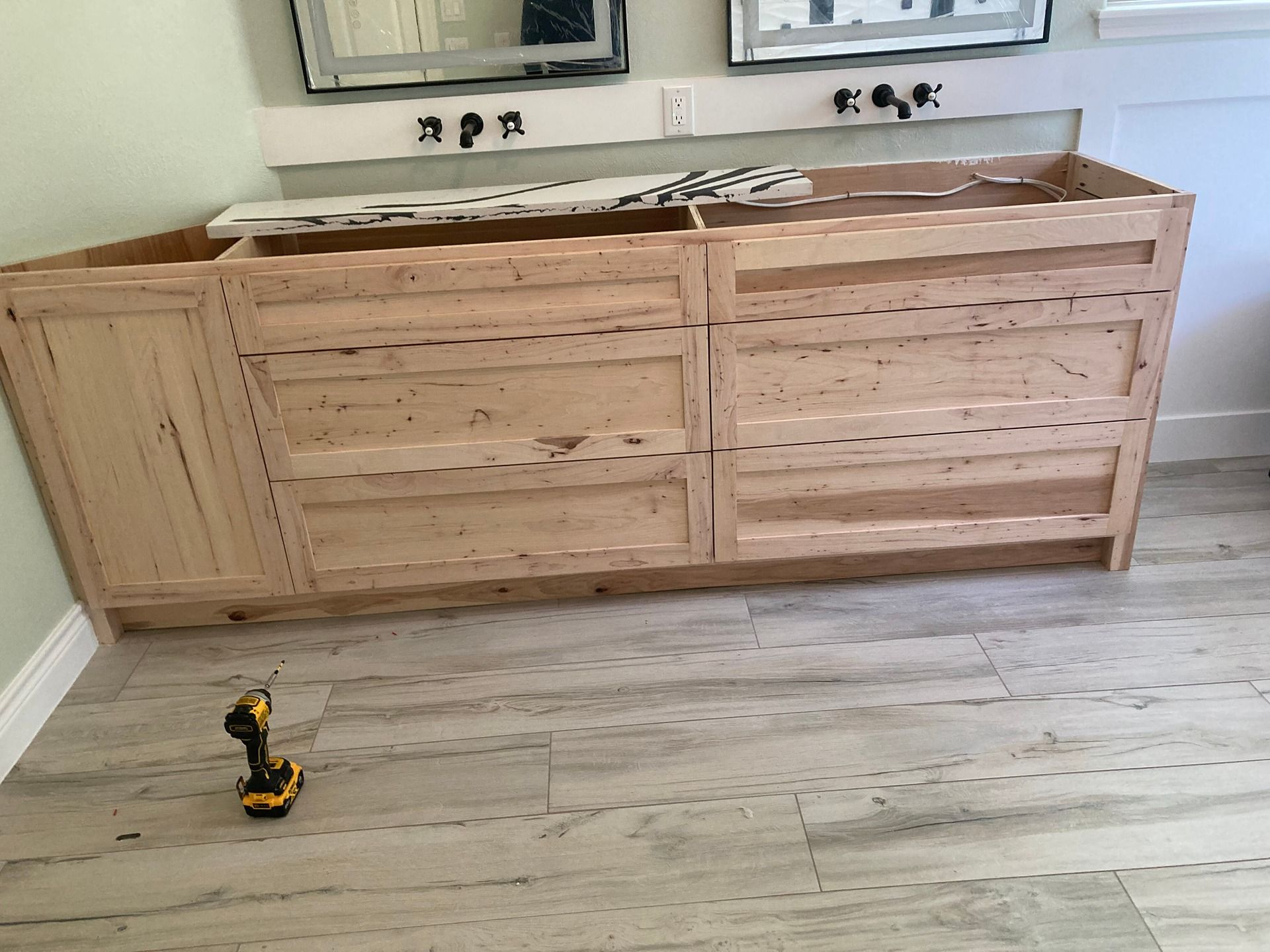 A raw wood double vanity base stands in a bathroom with light tile flooring and a cordless drill on the ground.