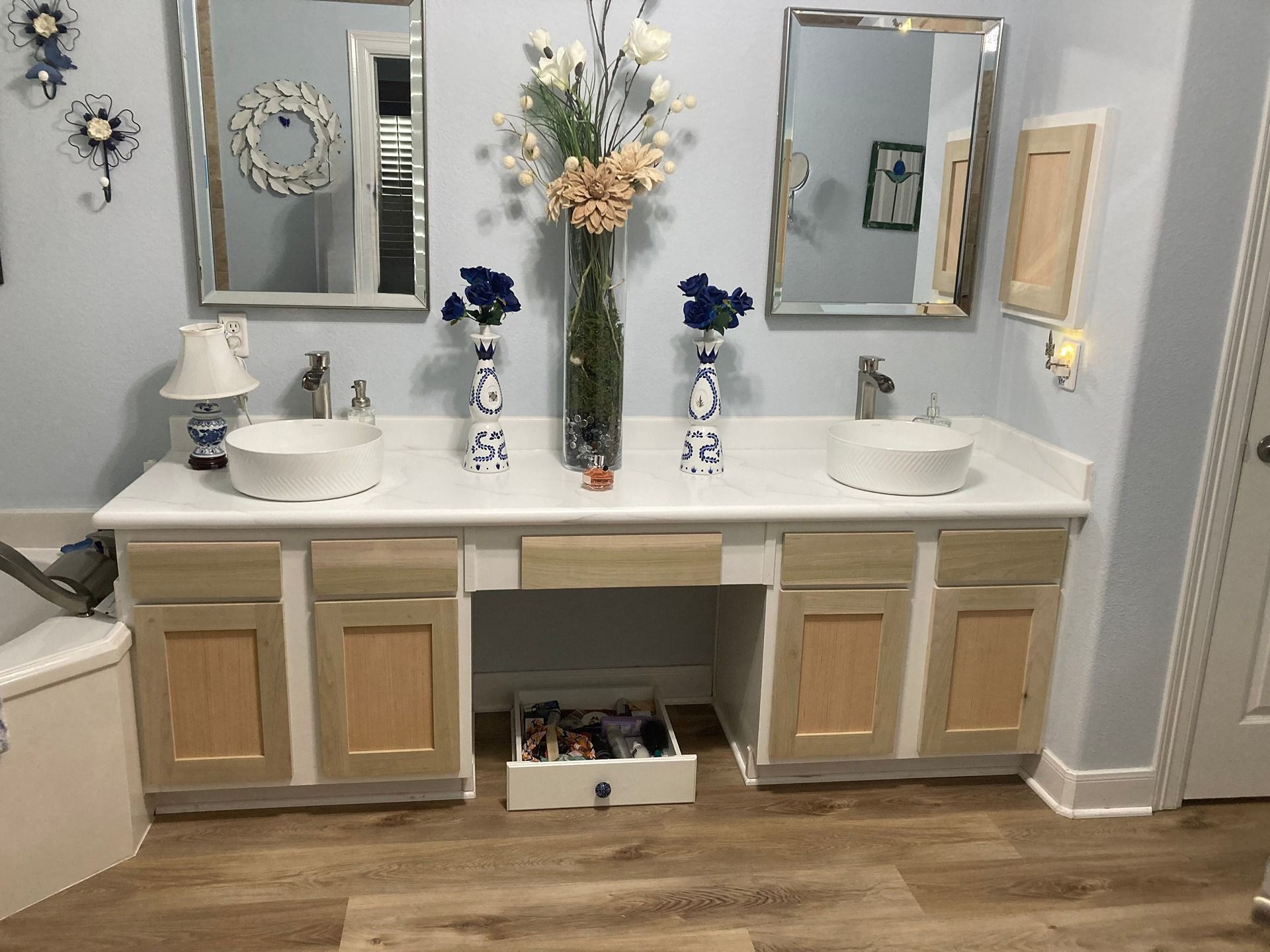 A double-sink bathroom vanity with wood-tone cabinet doors, white countertops, two mirrors, and floral decorations.