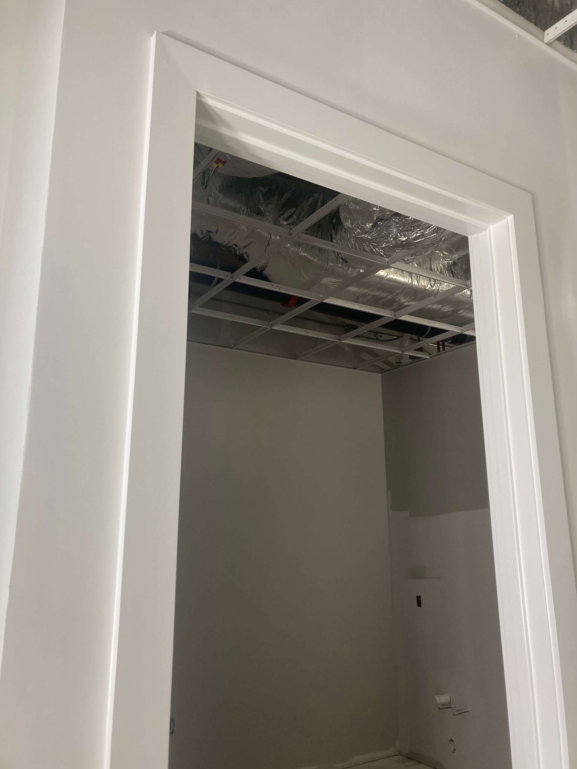 A white doorway frames a view into an unfinished room with exposed ceiling metal framing and gray painted walls.