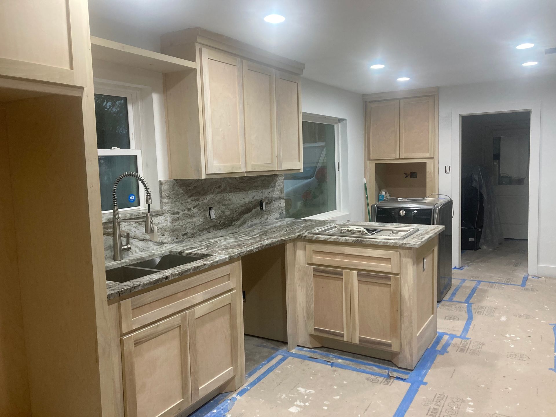 A kitchen under renovation with unfinished wood cabinets, granite countertops, and protective paper on the floor.
