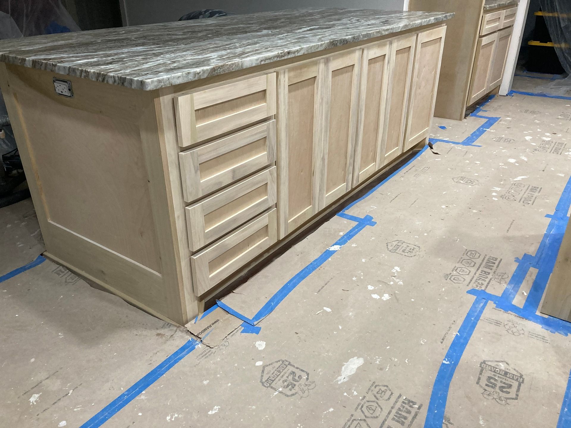 A light wood kitchen island with a granite countertop sits on a floor covered in protective paper and blue painter's tape.