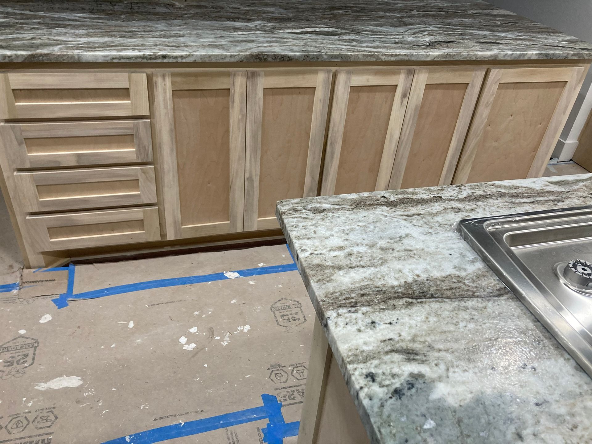 Unfinished wooden kitchen cabinets with a marble countertop in a room under construction with floor protection paper.