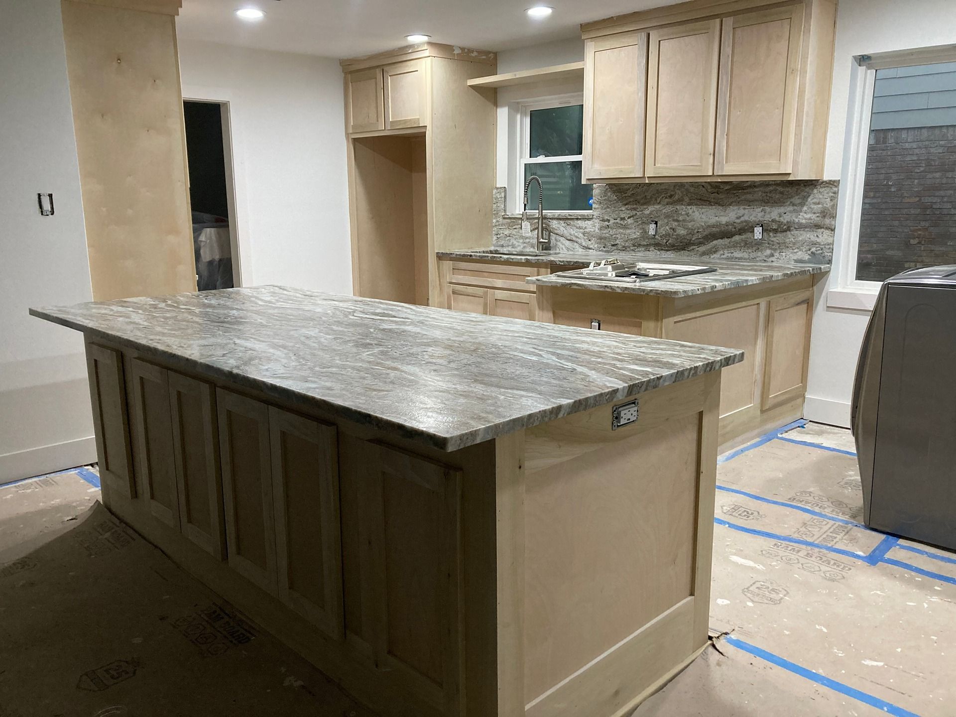 A kitchen under construction featuring a large island and counters with granite tops and light-colored wooden cabinets.