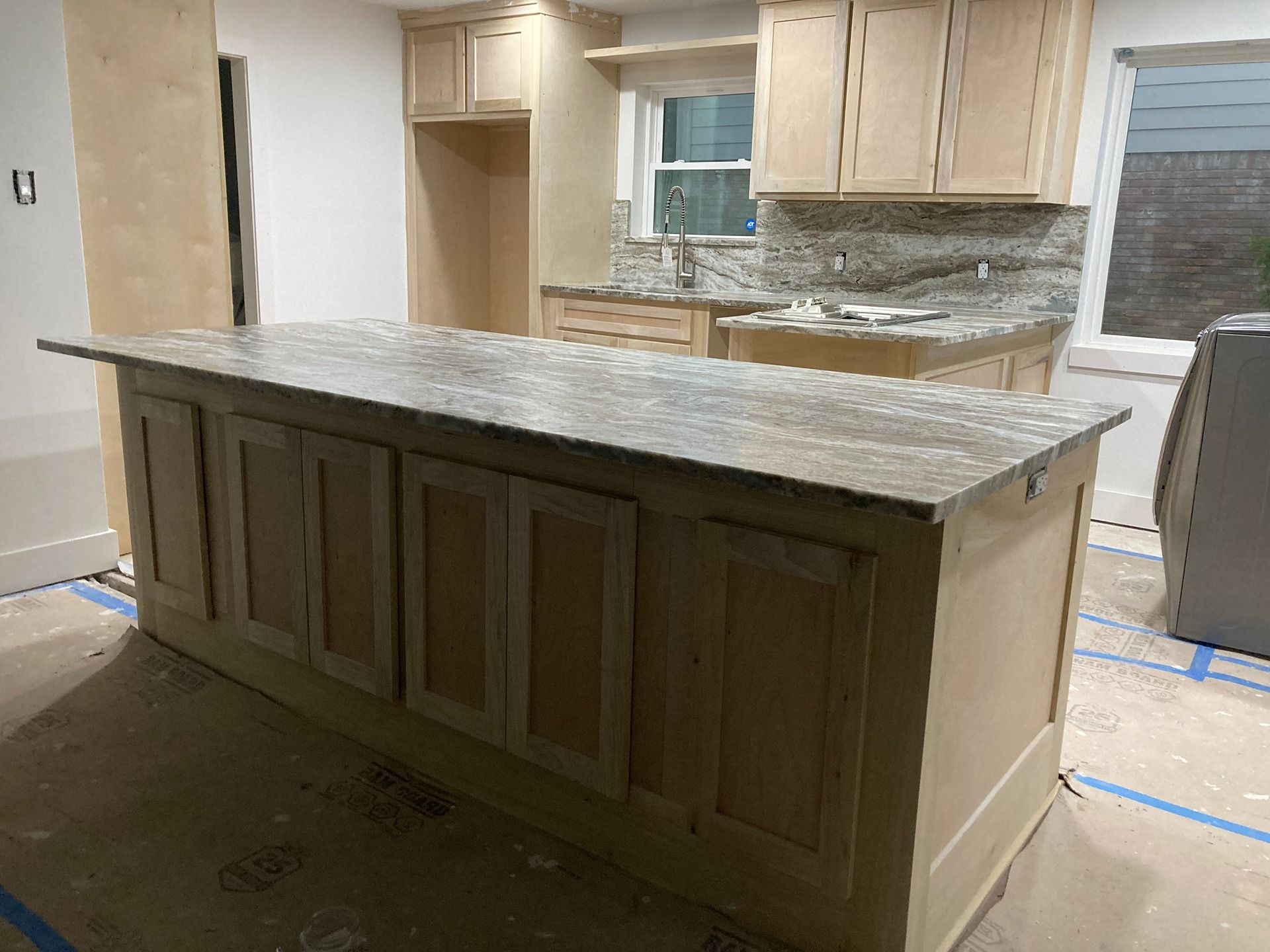 A kitchen island with a granite countertop sits in an unfinished room with light-colored cabinets and protective flooring.