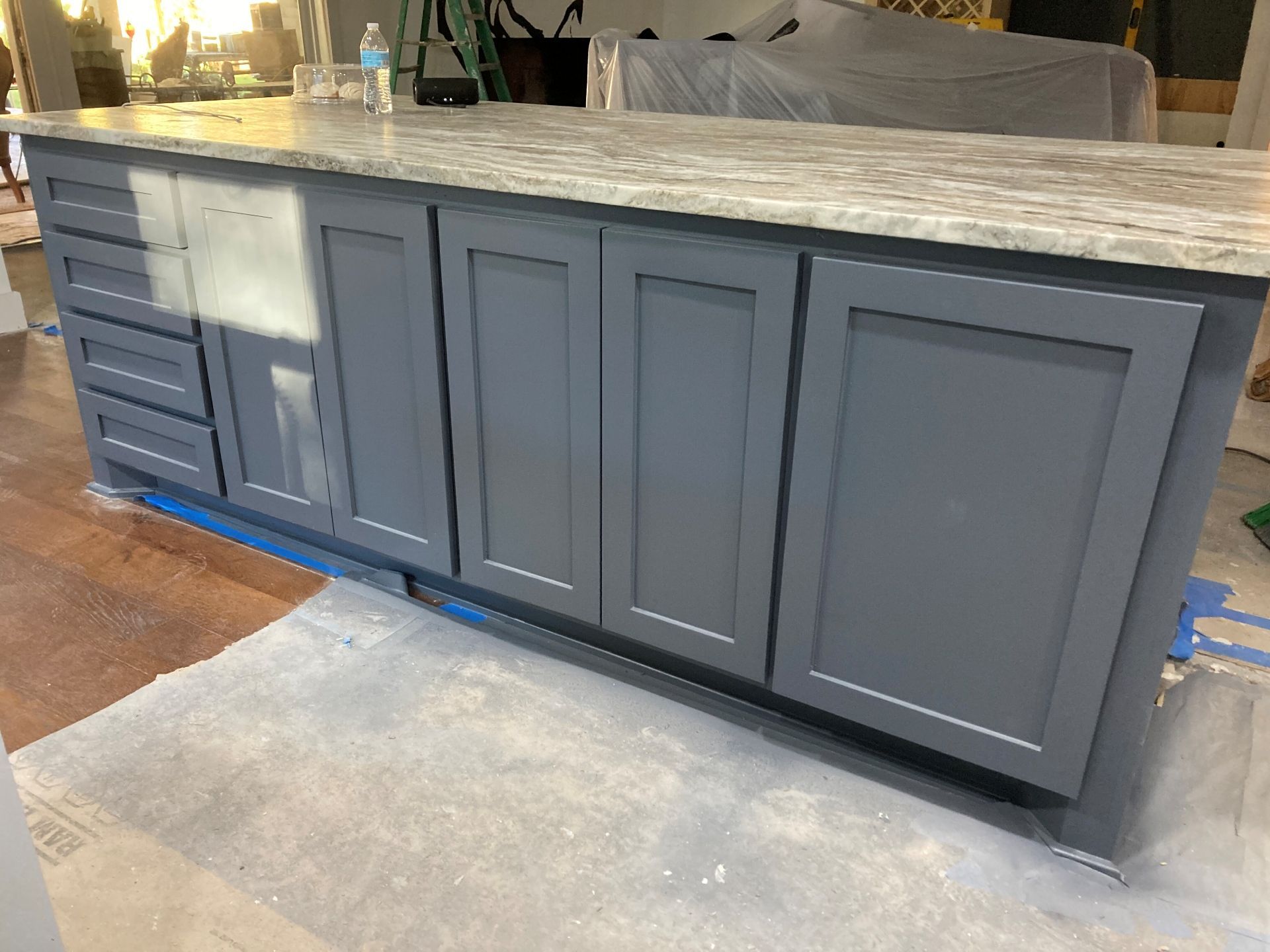 A long kitchen island with dark gray shaker-style cabinets and a light speckled countertop, set on a drop cloth.