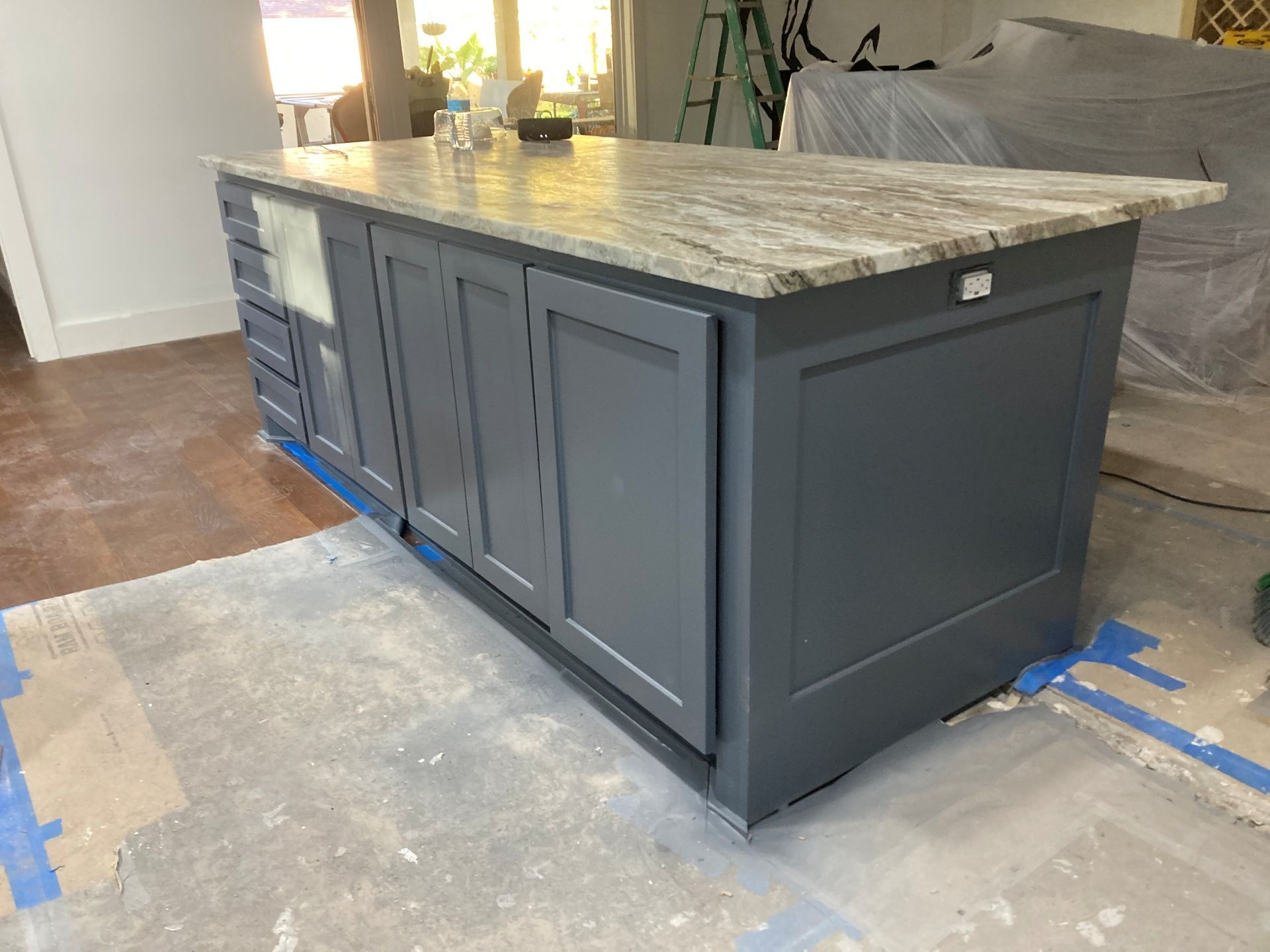 A dark gray kitchen island with a stone countertop sits on a floor protected by a plastic sheet during a renovation.