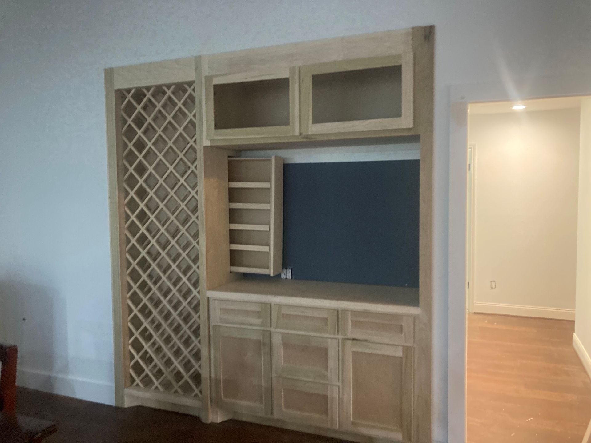 An unfinished light-wood built-in cabinet featuring a vertical wine rack, upper glass cabinets, shelving, and lower drawers.