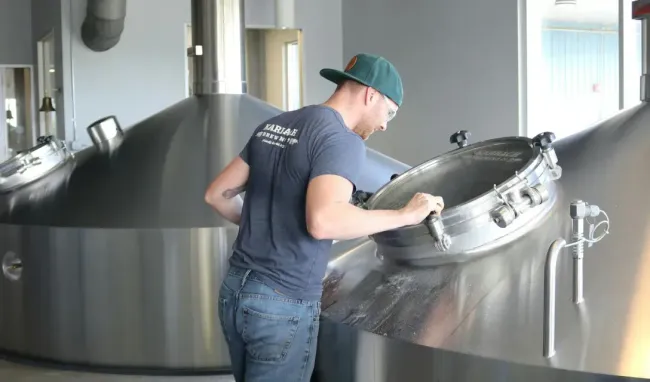 A man is standing next to a large metal tank in a brewery.