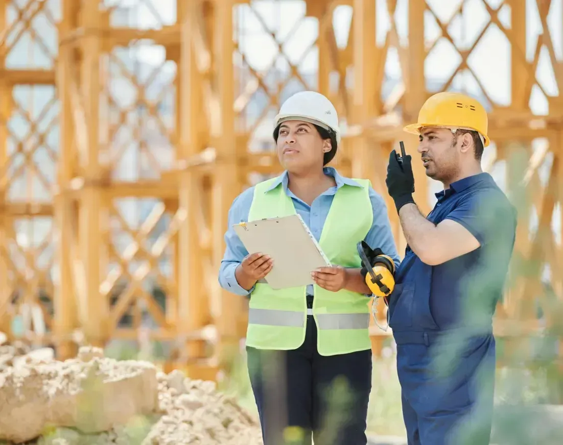 A man and a woman are standing on a construction site looking at a clipboard.