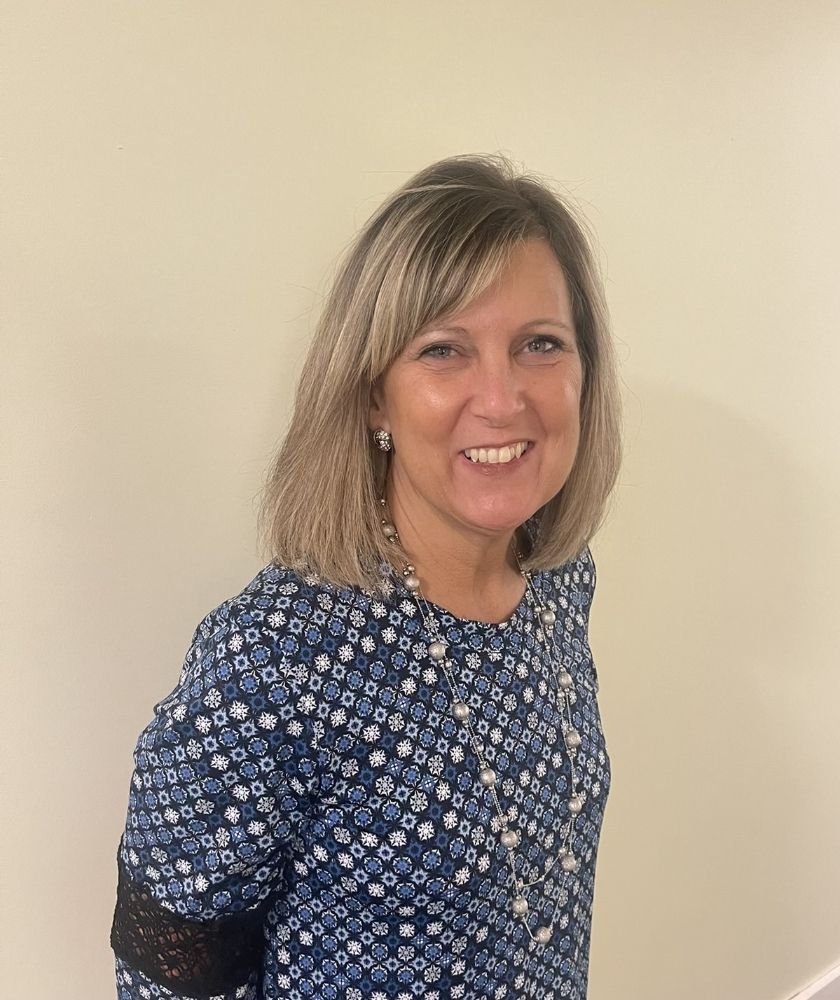 Penny Jones in a blue shirt is smiling and standing in front of a white wall.