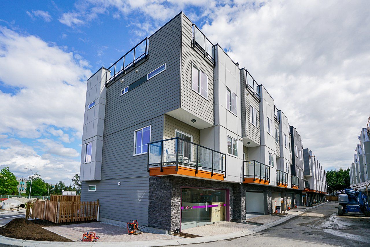 Modern gray and white townhouses with glass balconies and a cloudy sky overhead. A partially constructed building and a street are in front.