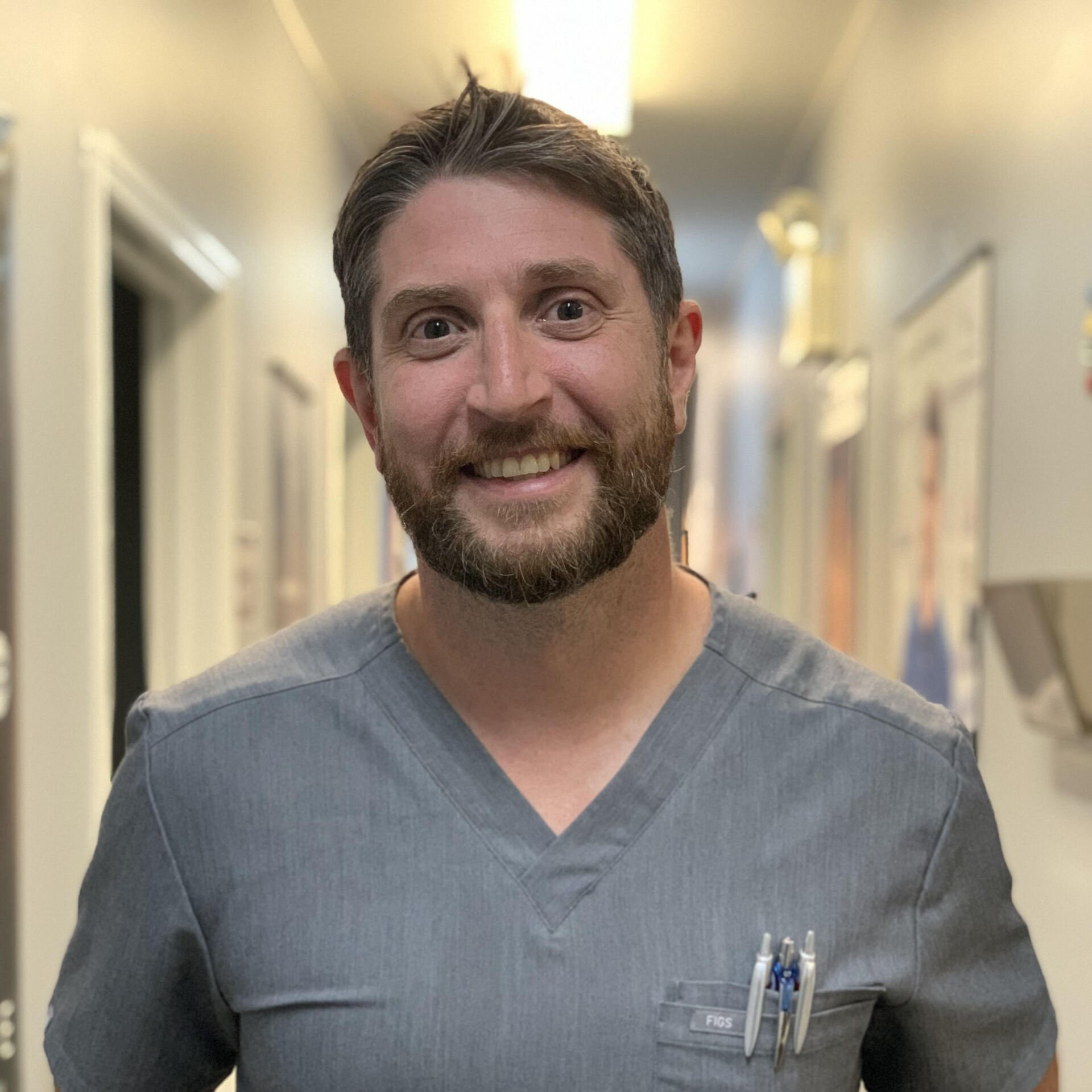 A man with a beard is wearing a scrub top and smiling in a hallway.