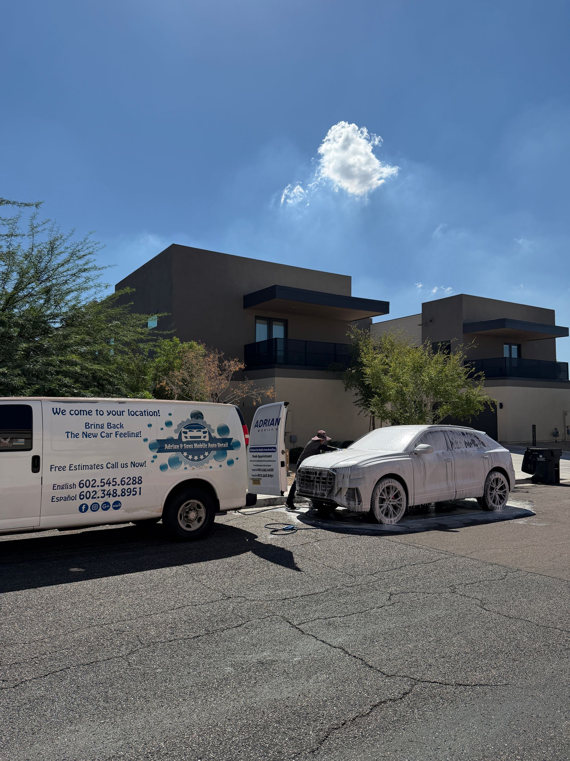 A white car covered in foam being washed next to a van. Buildings and a blue sky in the background.