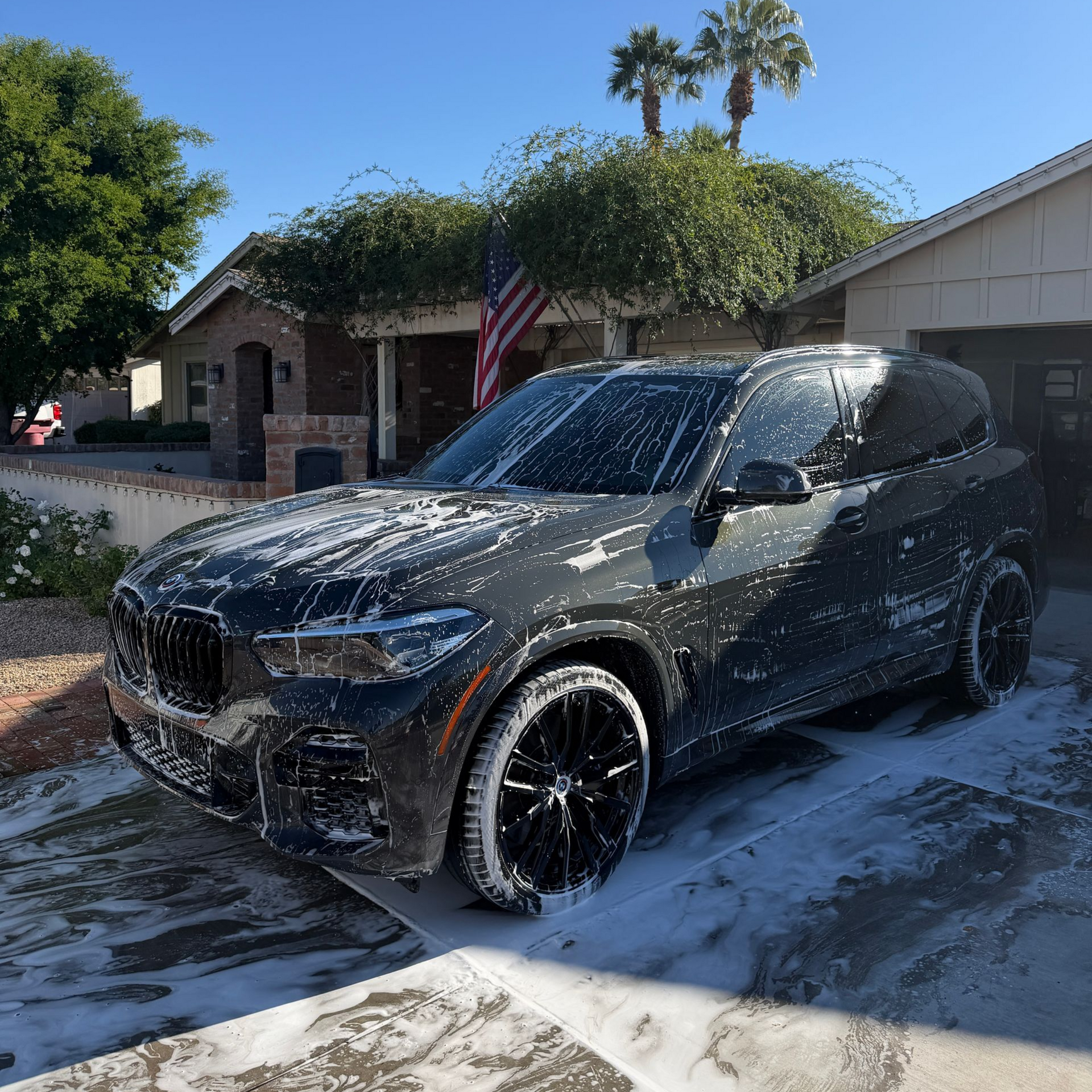 Dark gray SUV covered in white foam being washed in front of a house on a sunny day.