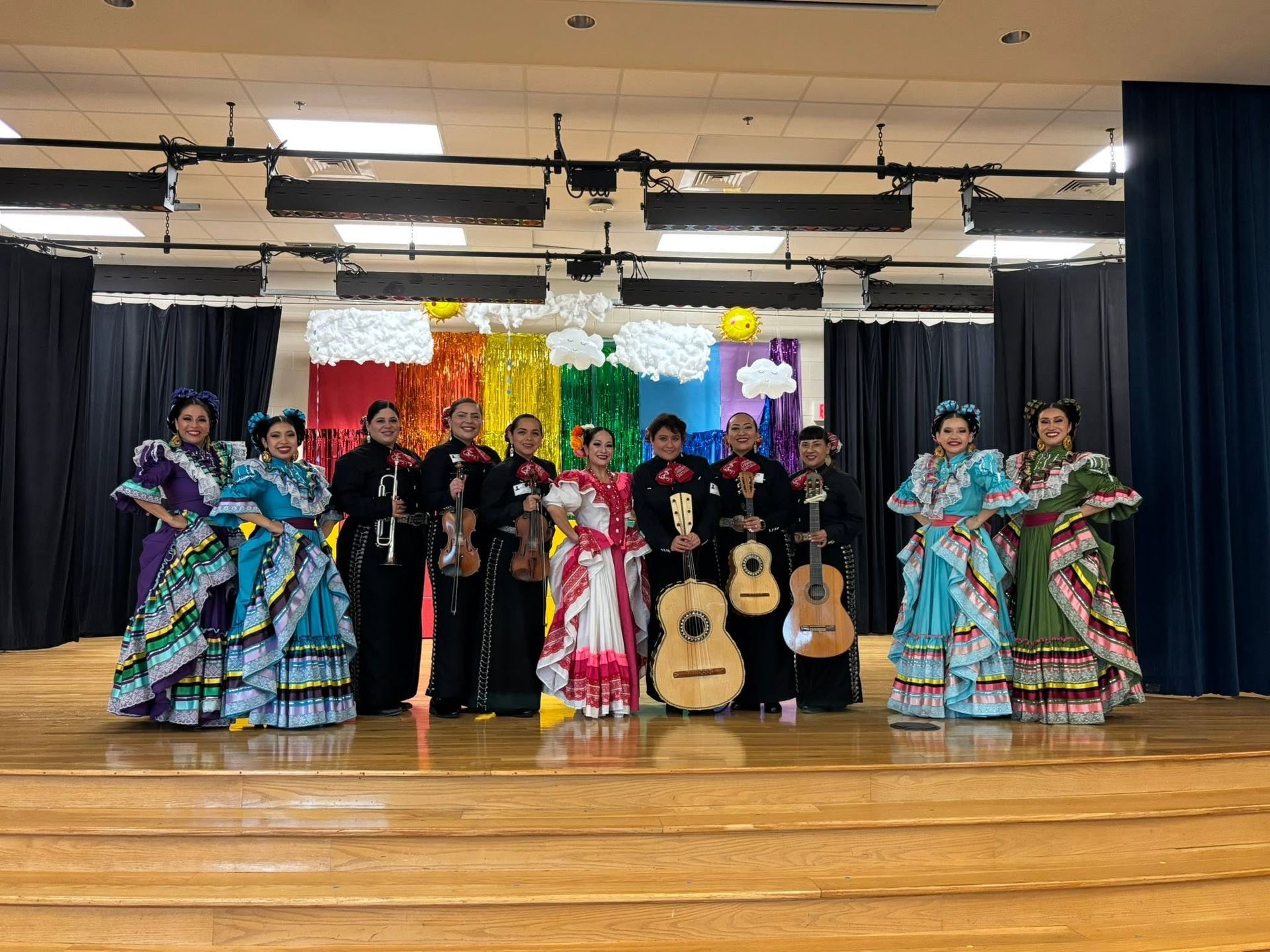 Mariachi band in black suits with colorful folkloric dancers on a stage with a black curtain backdrop