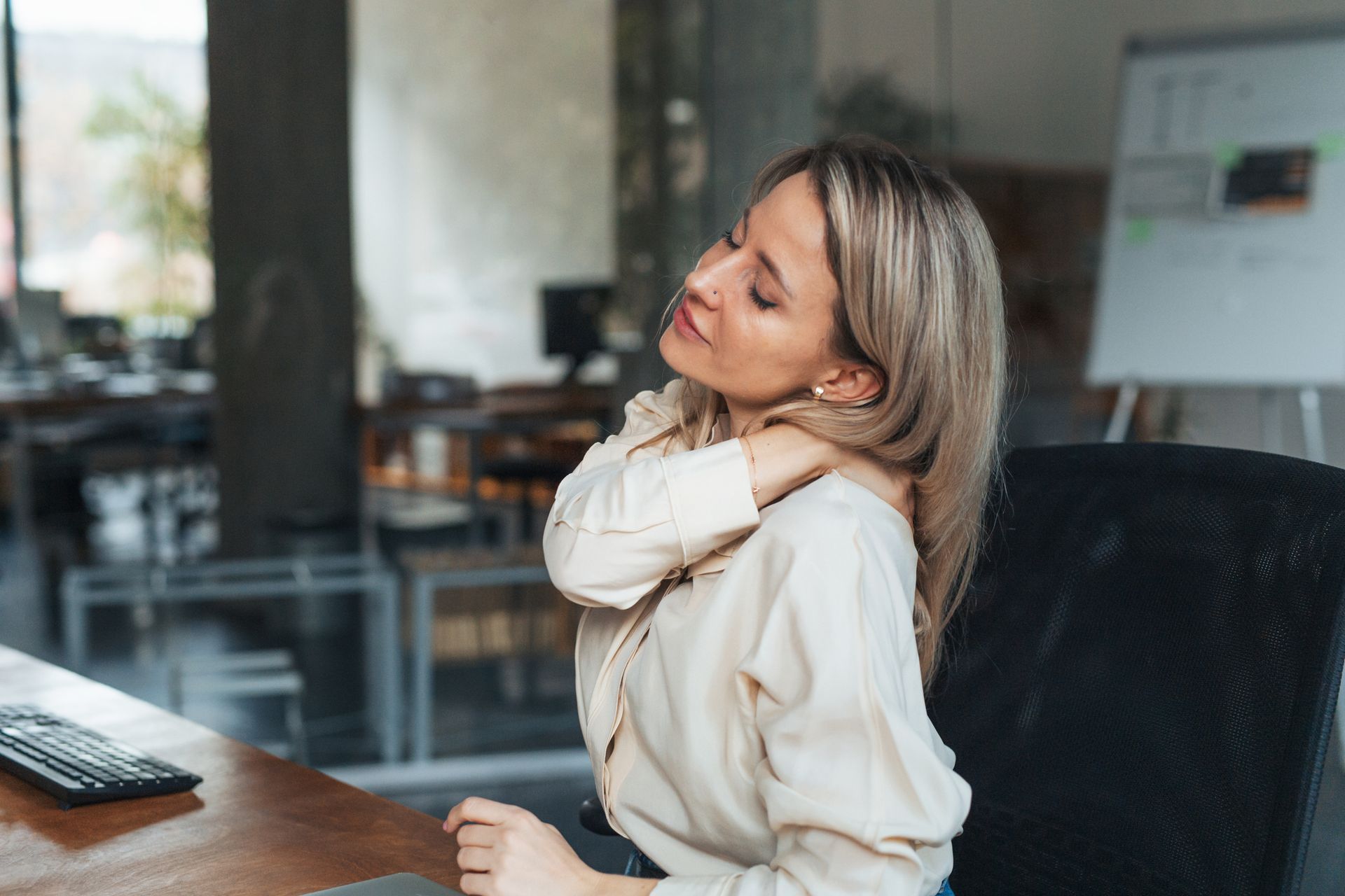 Woman in office experiencing neck pain, sitting at desk with computer and whiteboard.