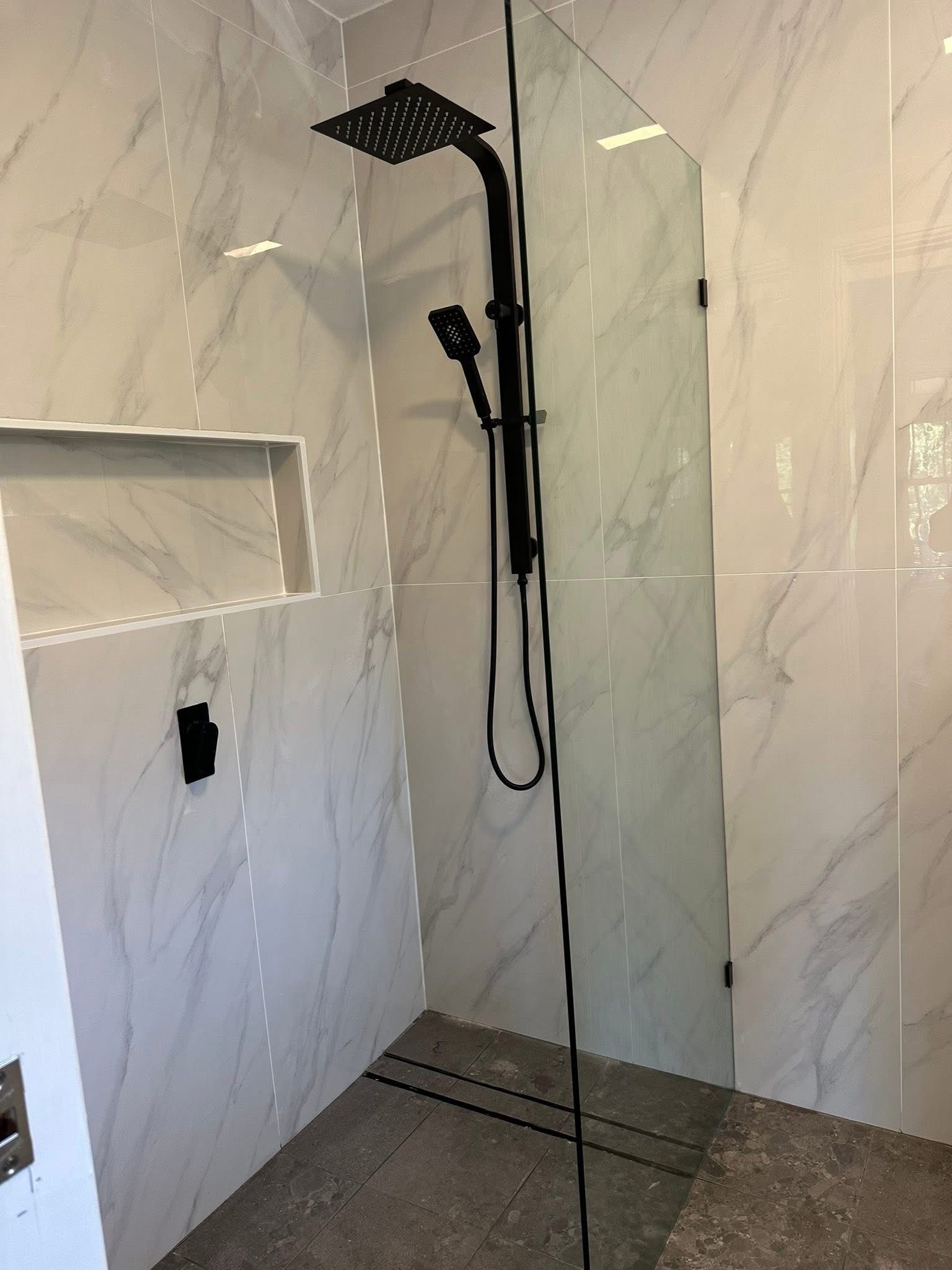 A shower with white marble-patterned tile, a black rainfall showerhead, a handheld sprayer, and a glass partition.