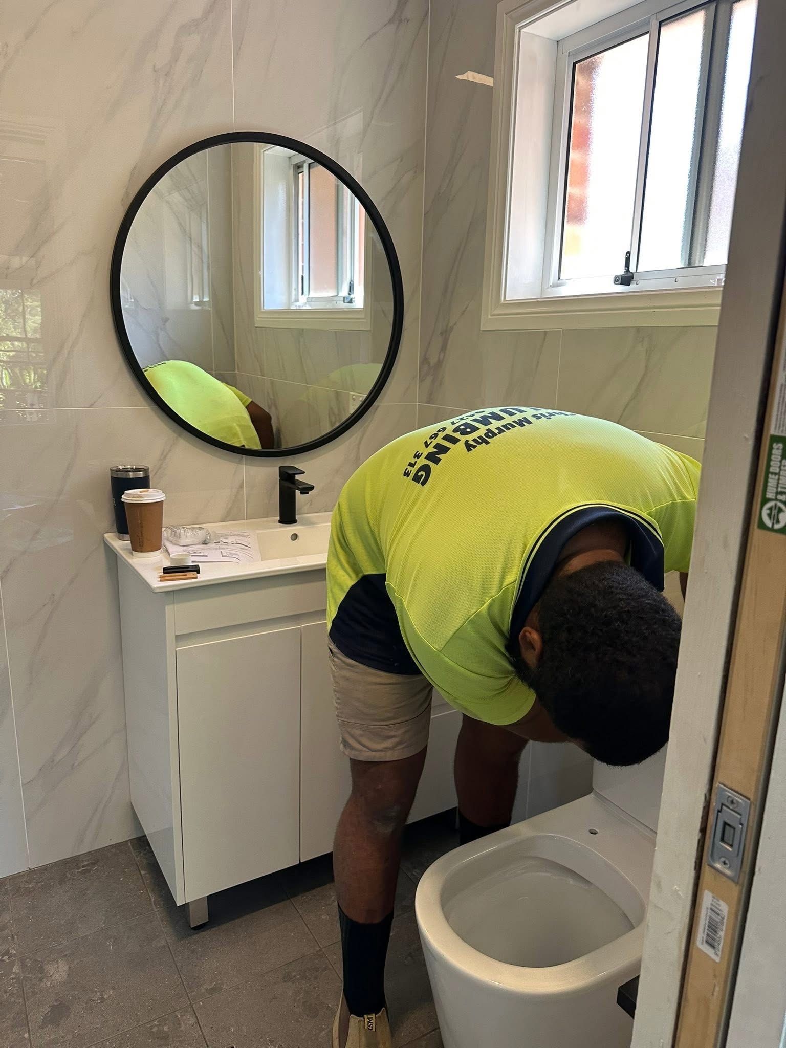 A person in a high-visibility yellow shirt is installing a toilet in a bathroom with white marble-patterned walls.