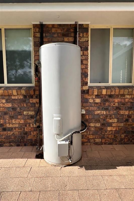A large grey cylindrical water heater sits on a paved patio against a brick wall between two windows.