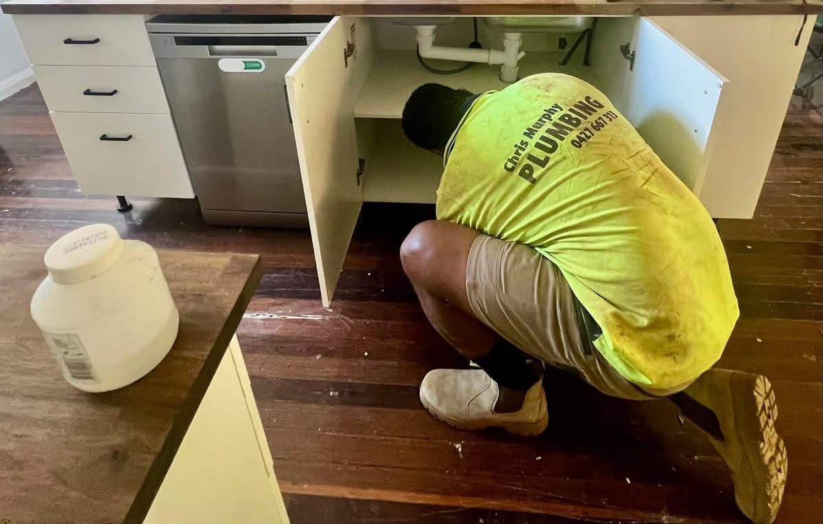 A plumber wearing a high-visibility yellow shirt works on pipes inside an open kitchen cabinet.