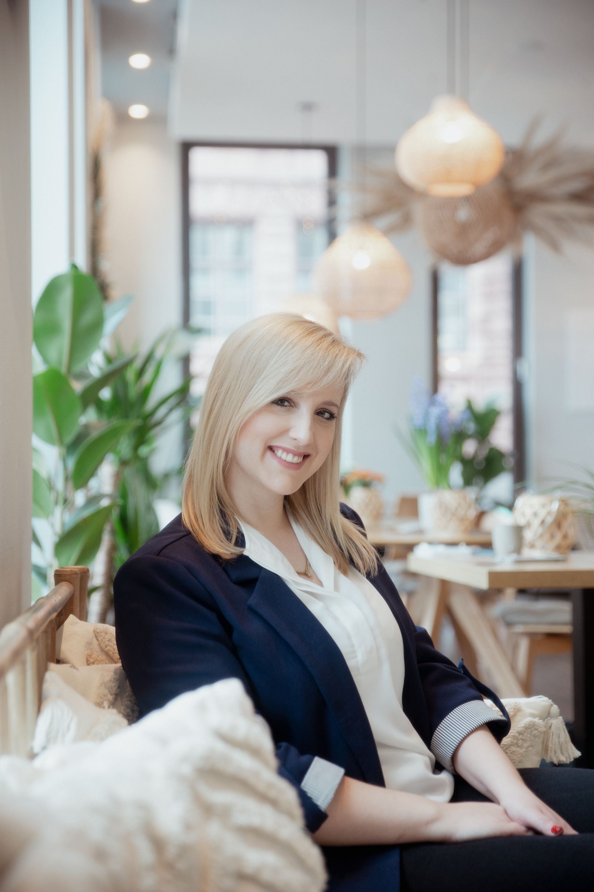 Blonde Frau im Blazer lächelt, sitzt drinnen. Neutrale Töne, hereinströmendes Licht.