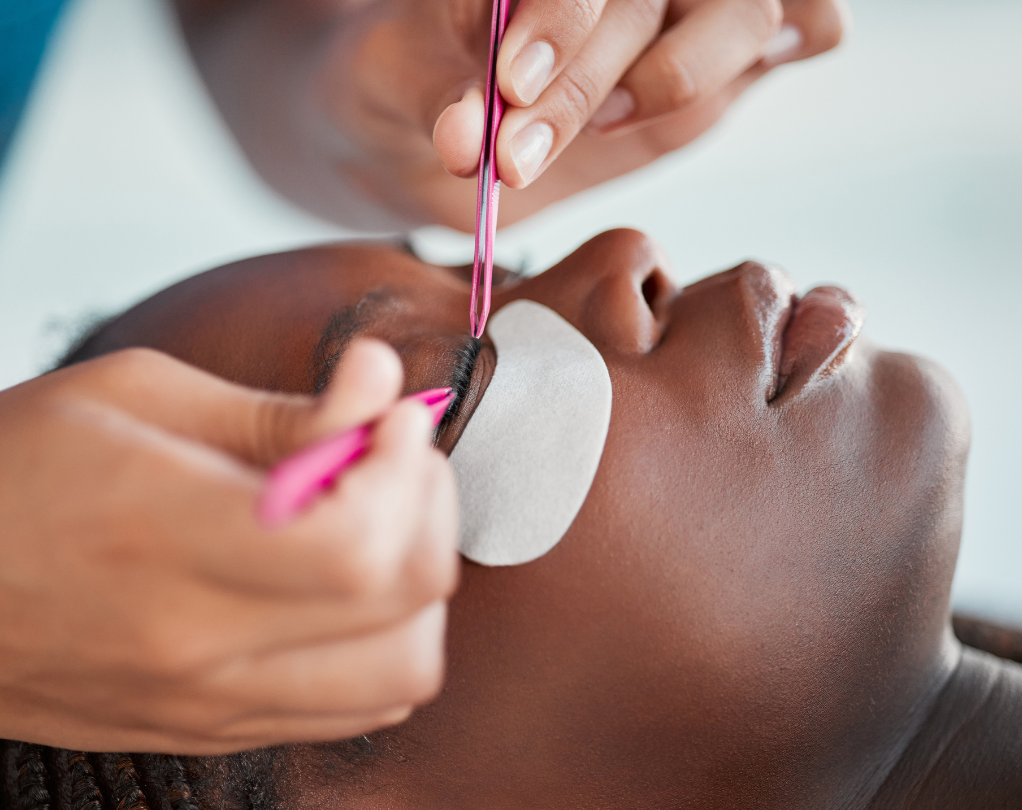 A person receiving eyelash extensions. A technician uses pink tweezers to apply lashes to the closed eye, with a white eye patch in place.