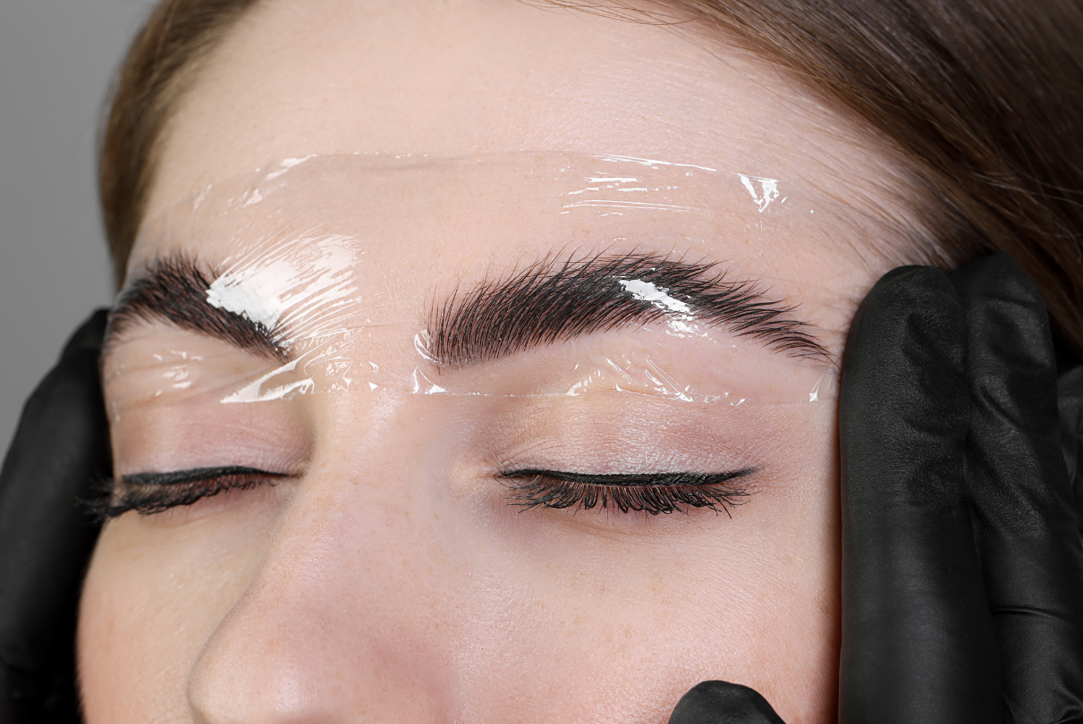 Woman with dark eyebrows, eyes closed, receiving a brow lamination treatment. A clear gel is applied. Hands in black gloves frame her face.