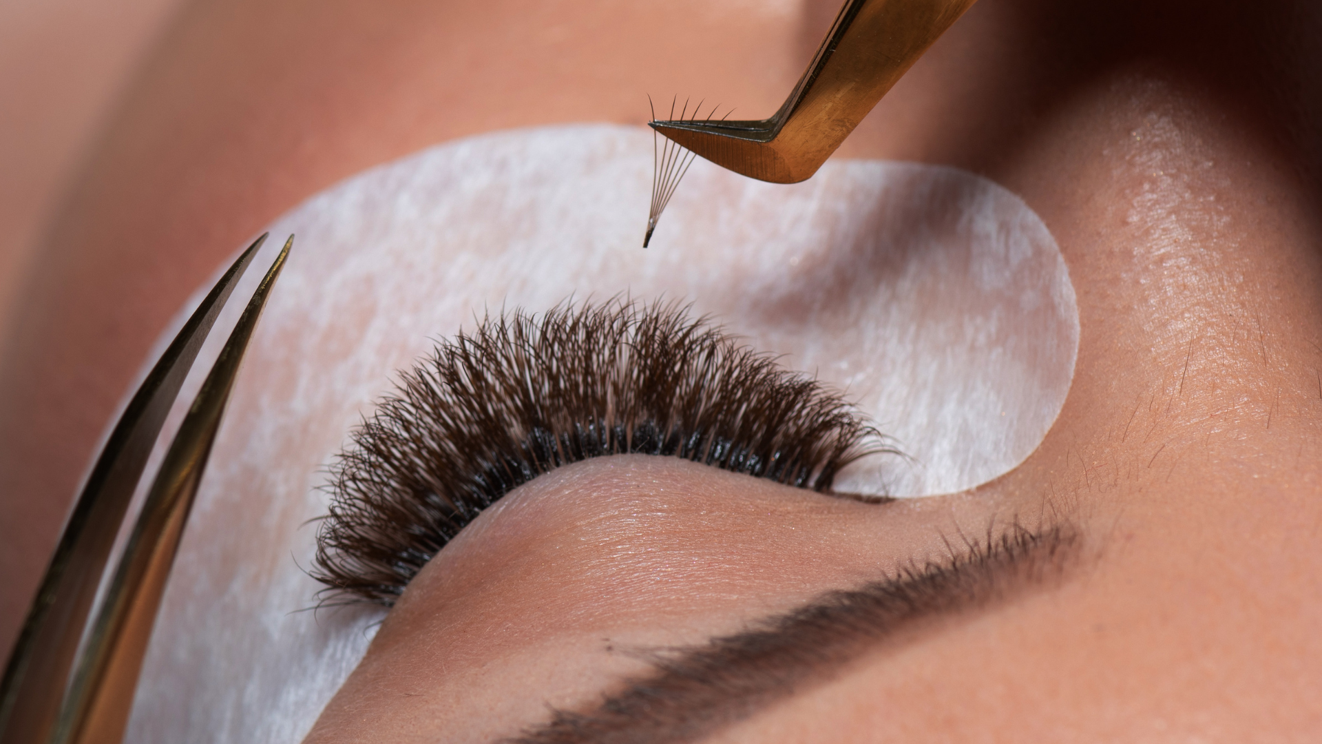 A person receiving eyelash extensions. A hand holds tweezers, picking up a lash from a tray while another hand holds the tray.
