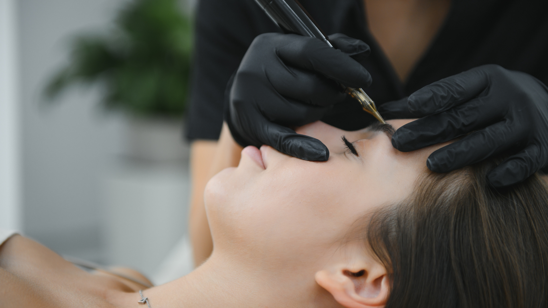 Person receiving an eyebrow microblading procedure. A technician in black gloves uses a handheld device on the woman's face.