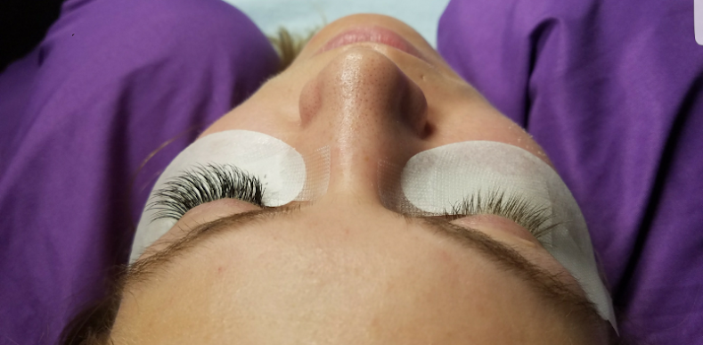 A woman with newly applied eyelash extensions, lying down with under-eye pads. She has purple clothing and is in a beauty salon.