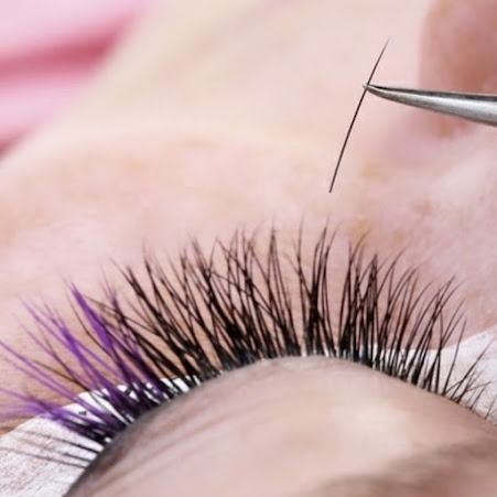 Close-up of someone receiving a lash extension. Black and purple lashes are visible, with tweezers holding an individual lash.