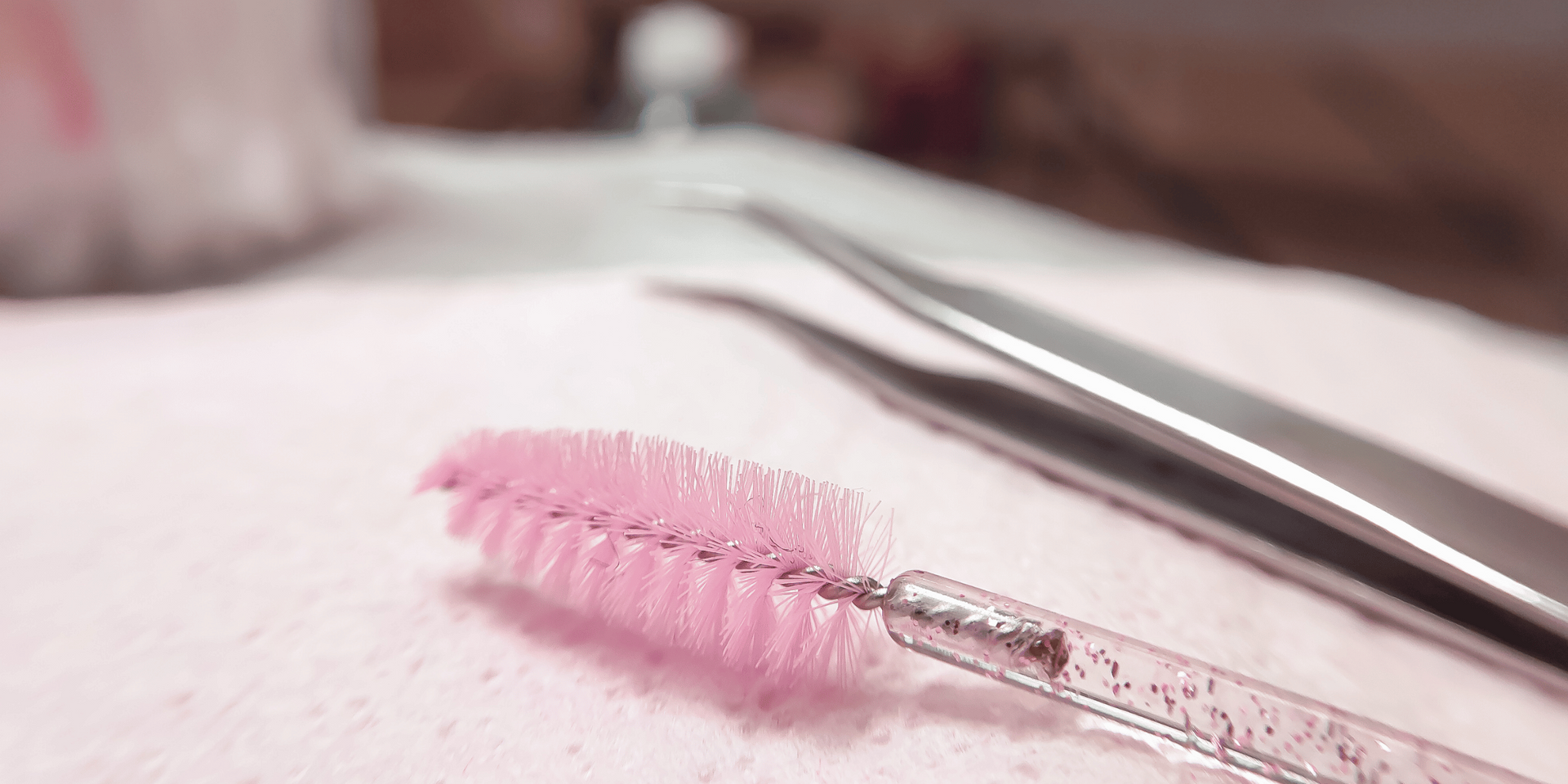Pink mascara wand next to silver tweezers on a white surface.