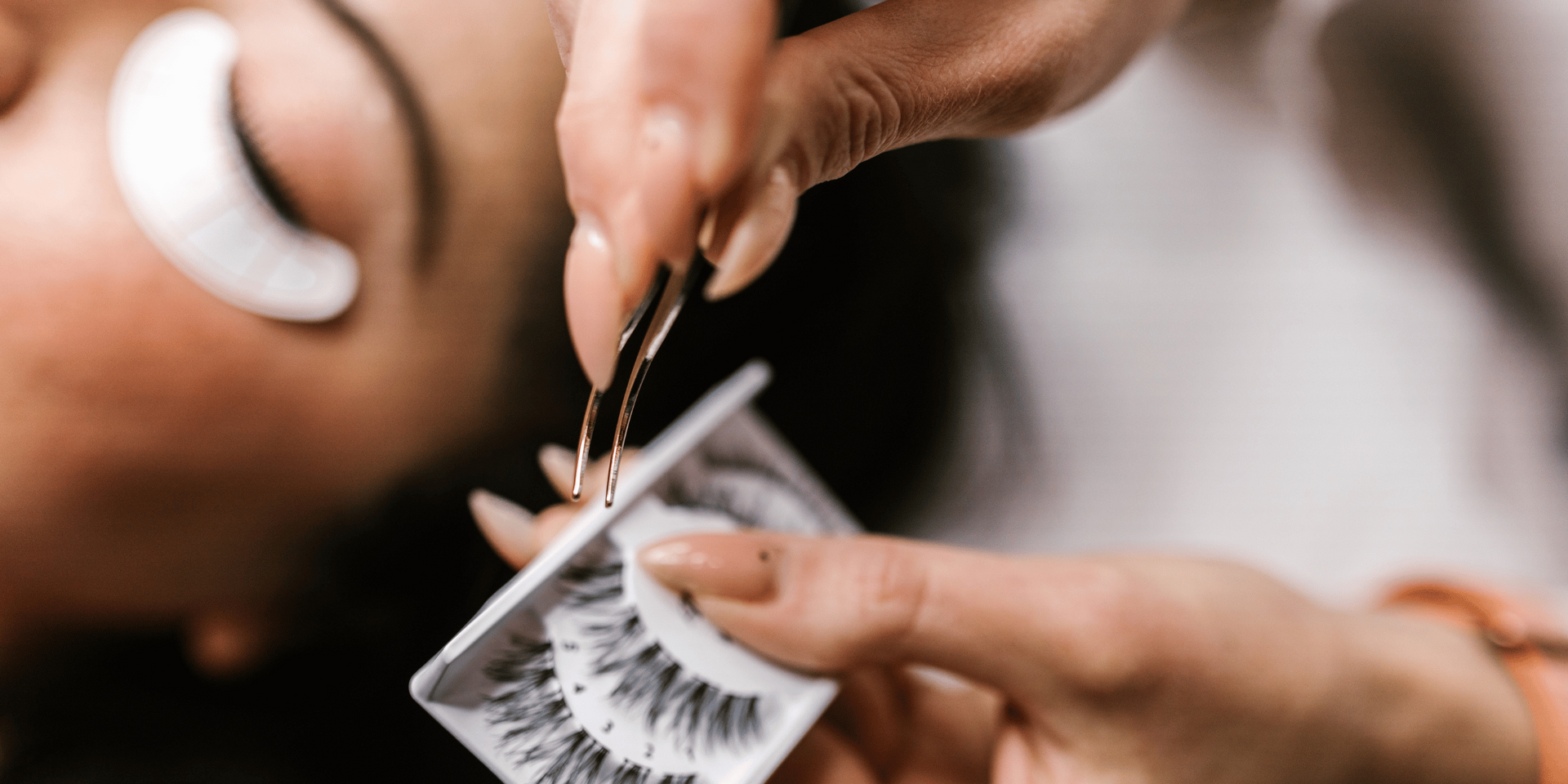 A person's eye with a white patch is getting eyelash extensions applied. A hand holds tweezers and a tray of false eyelashes.