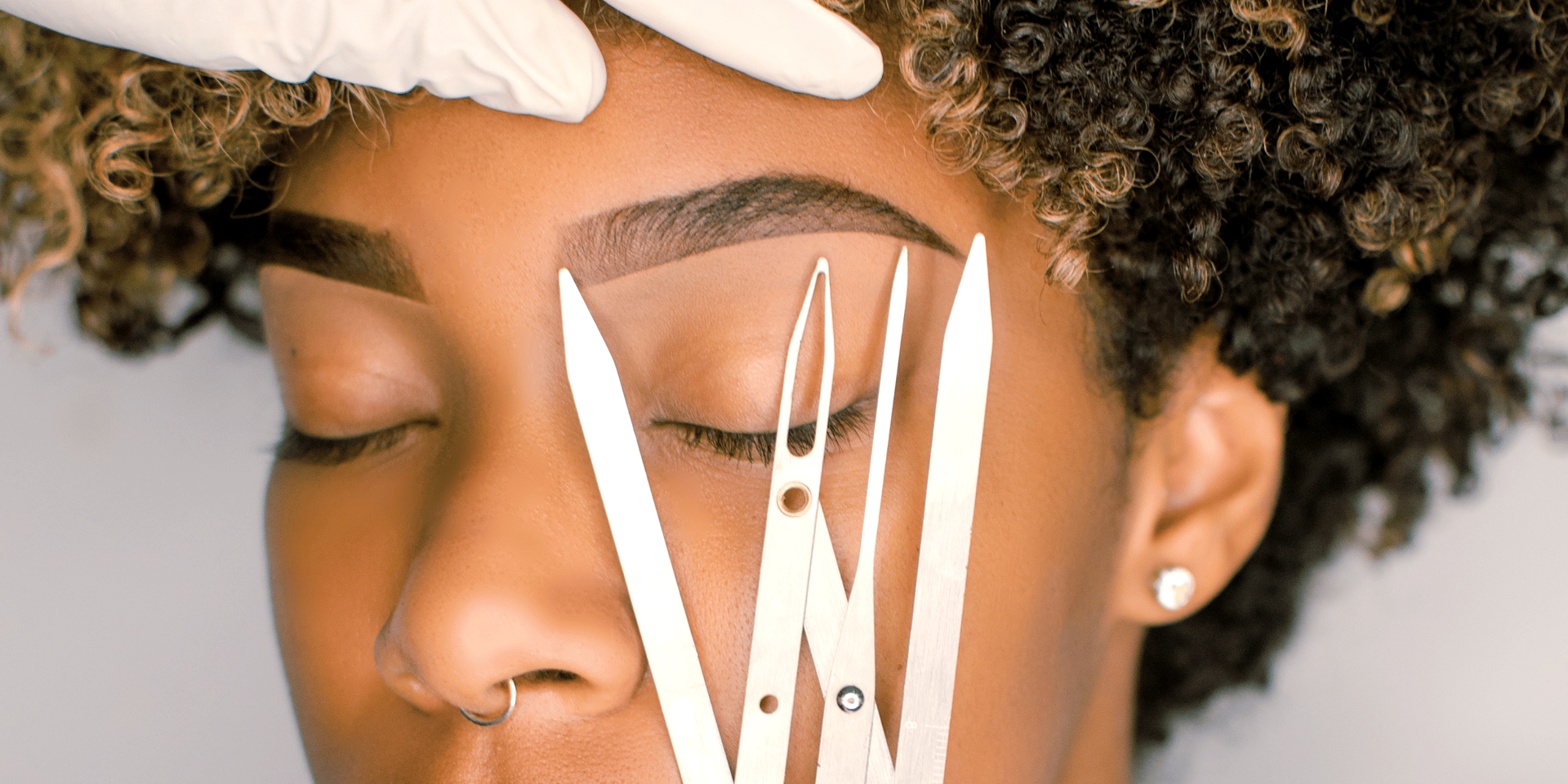 A person getting their eyebrows measured for a shaping procedure. A gloved hand holds instruments to the model's closed eye.
