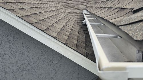 Roof edge with a white gutter and ladder leaning against gray shingles