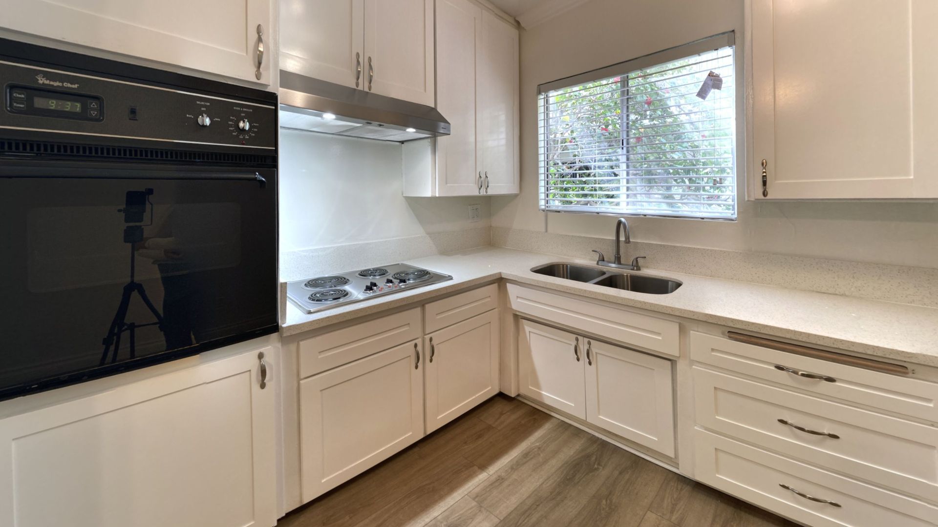 White kitchen with oven, stovetop, sink, cabinets, window, and wood-look flooring.