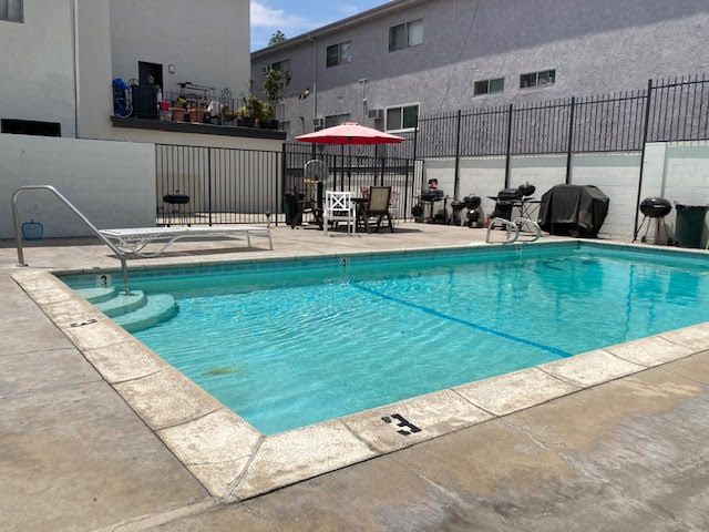 Outdoor pool with a red umbrella, lounge chair, and barbecue grills, surrounded by a fence and apartment buildings.