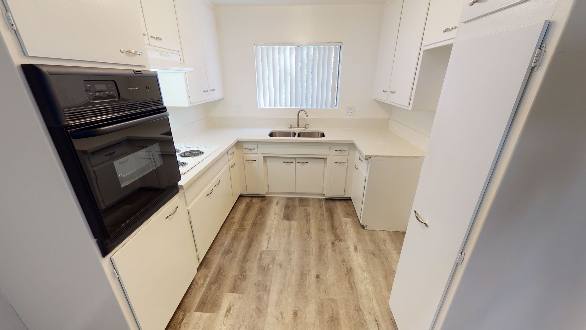 Kitchen with white cabinets, appliances, and light wood-look flooring. Window with blinds.