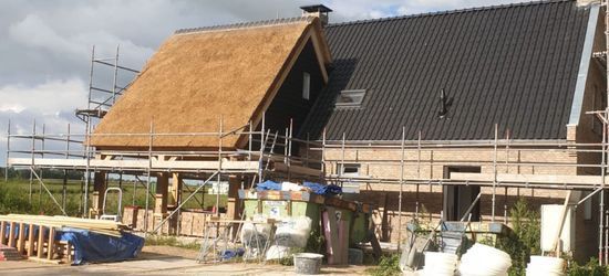Een huis in aanbouw met een rieten dak aan de ene kant en zwarte dakpannen aan de andere kant, omgeven door steigers.