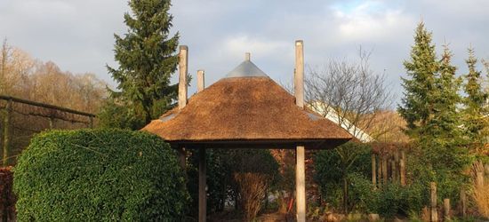 Prieel met rieten dak in een tuin, omgeven door bomen en struiken onder een bewolkte hemel.