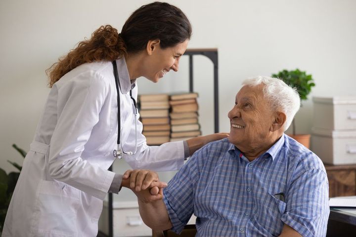 Doctor in white coat smiling, holding the hand and touching the shoulder of a seated older person in a blue shirt.