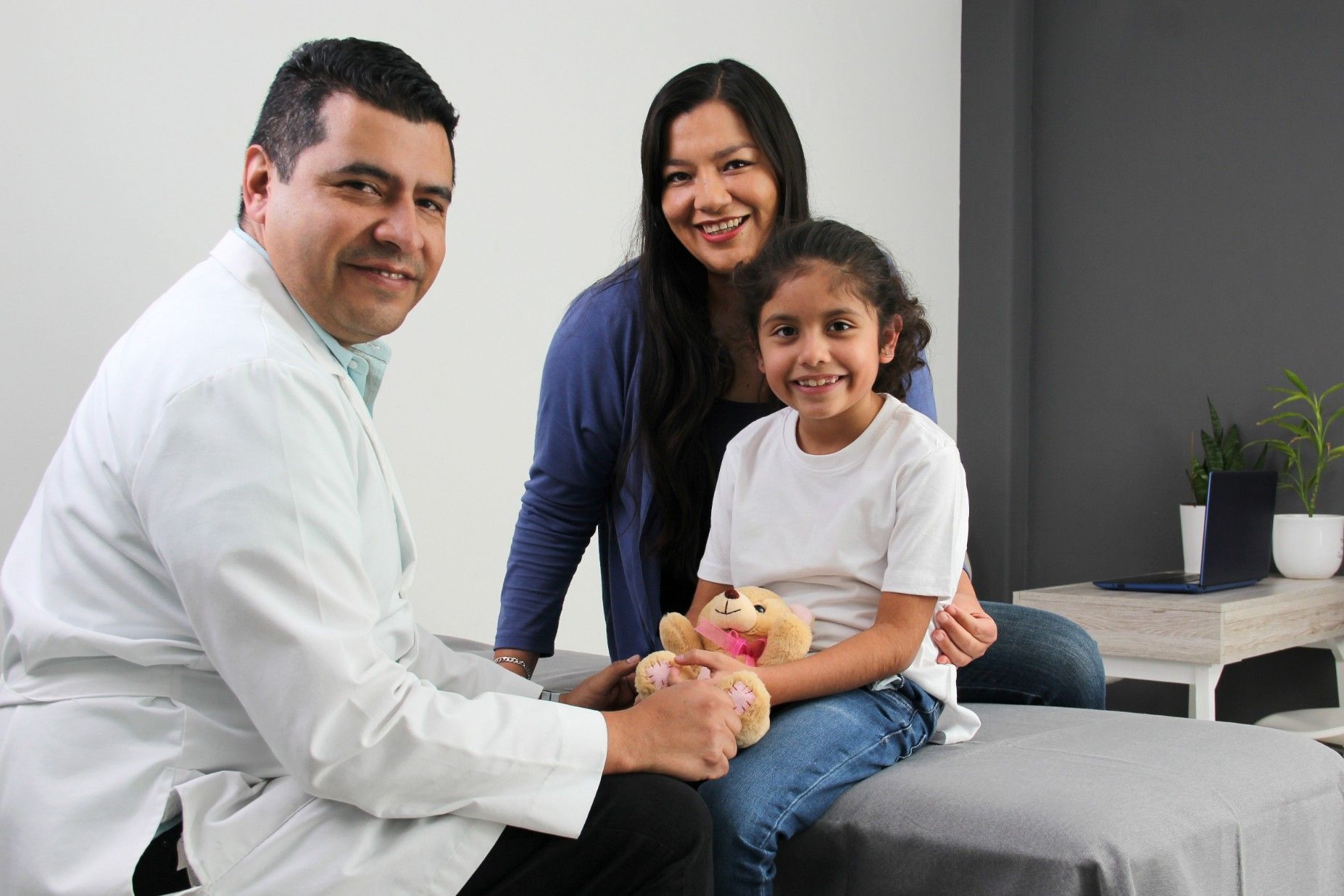 Doctor with a child and parent; doctor smiles, child holds toy, parent smiles; office setting.