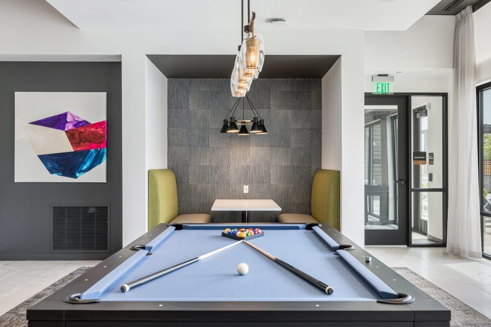 Modern apartment common area with a blue felt pool table set up for a game, geometric wallpaper, abstract wall art, stylish pendant lights, and a green chair. Glass doors lead to another room, and an exit sign is visible above the doorway at Marq Perimeter in Atlanta, GA.