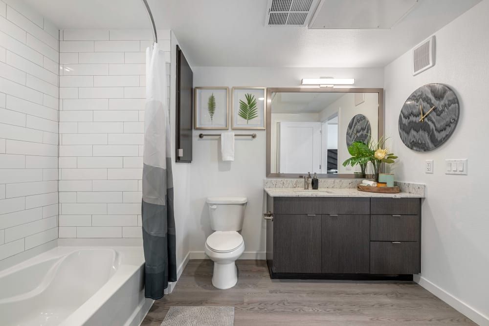 A modern bathroom interior with white subway tiled walls, a gray shower curtain on the left covering a bathtub, and a dark wood vanity with a marble countertop on the right featuring a basin sink. Above the vanity, a large mirror reflects the bathroom's interior, flanked by wall art depicting ferns and a decorative clock with a marbled design. The floor is covered with gray wood-like tiles, and a textured gray mat lies in front of the vanity at Marq Perimeter in Atlanta, GA.