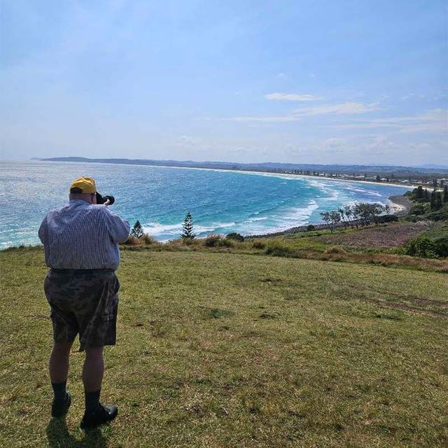 a man is standing on top of a grassy hill and taking a photo overlooking the ocean .