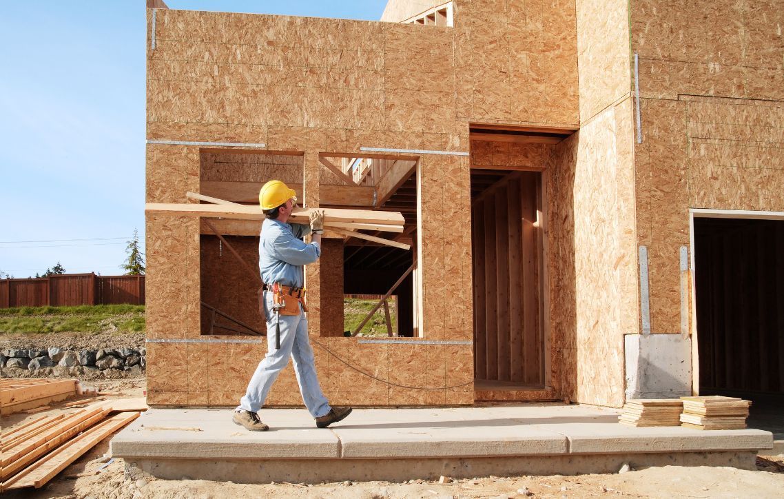 A construction worker is carrying a piece of wood in front of a house under construction.