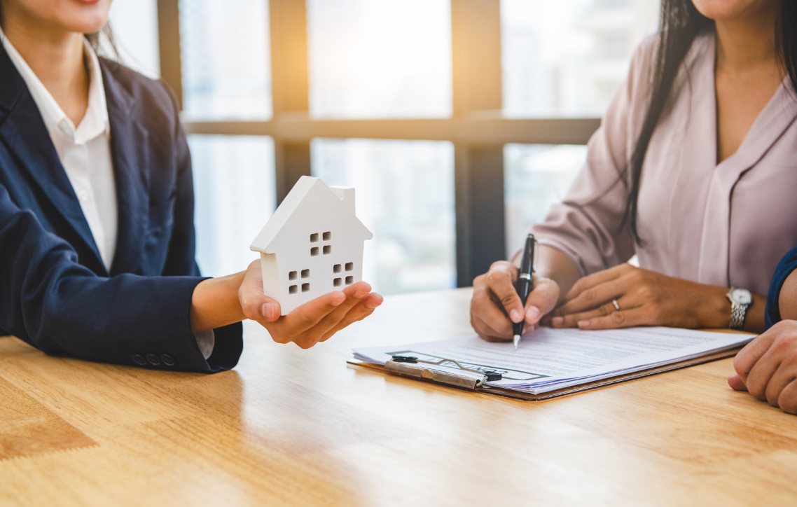A woman is holding a model house while another woman is signing a document.